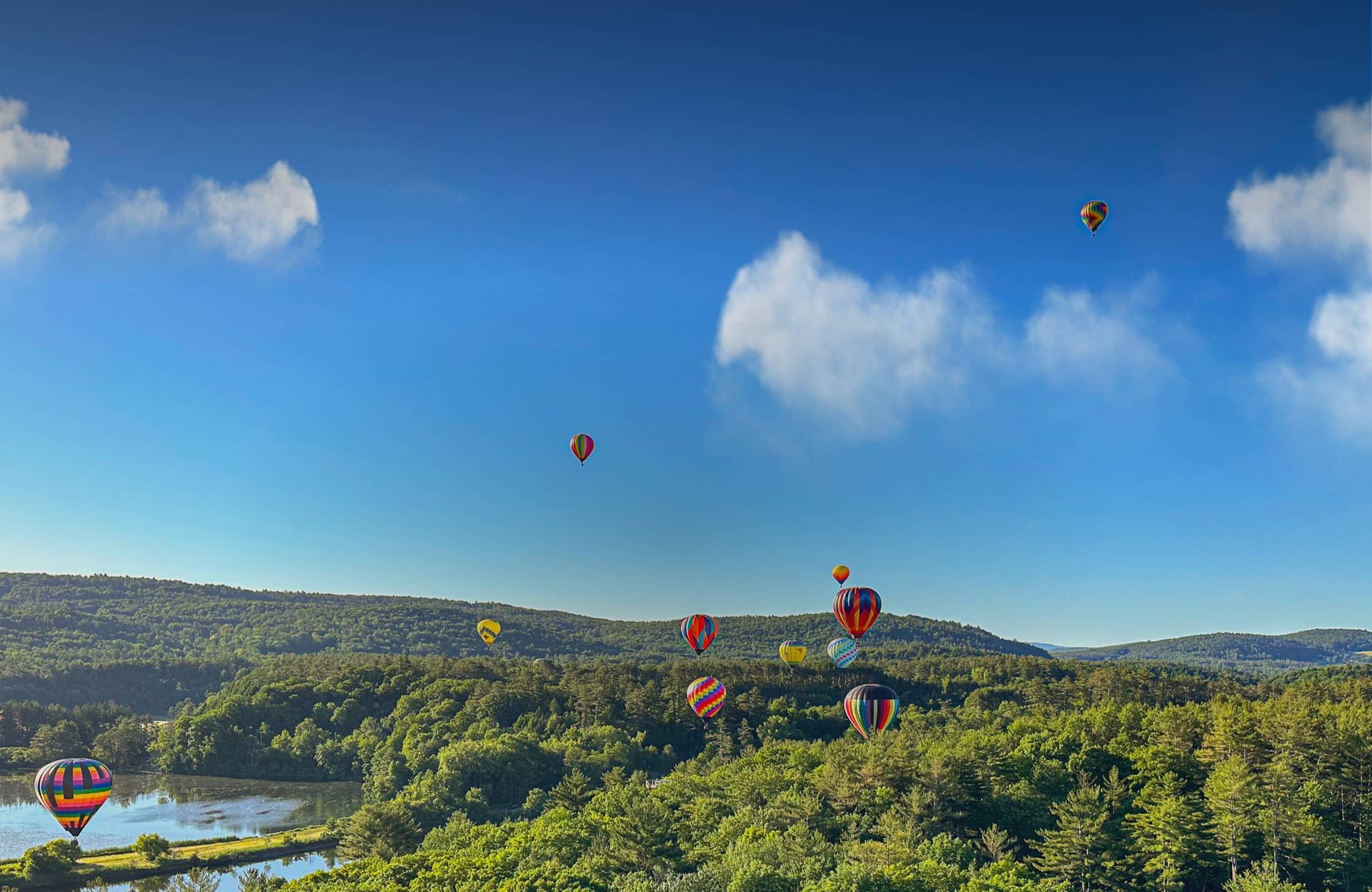 Multiple hot air balloons floating above a forested landscape with a lake and blue sky